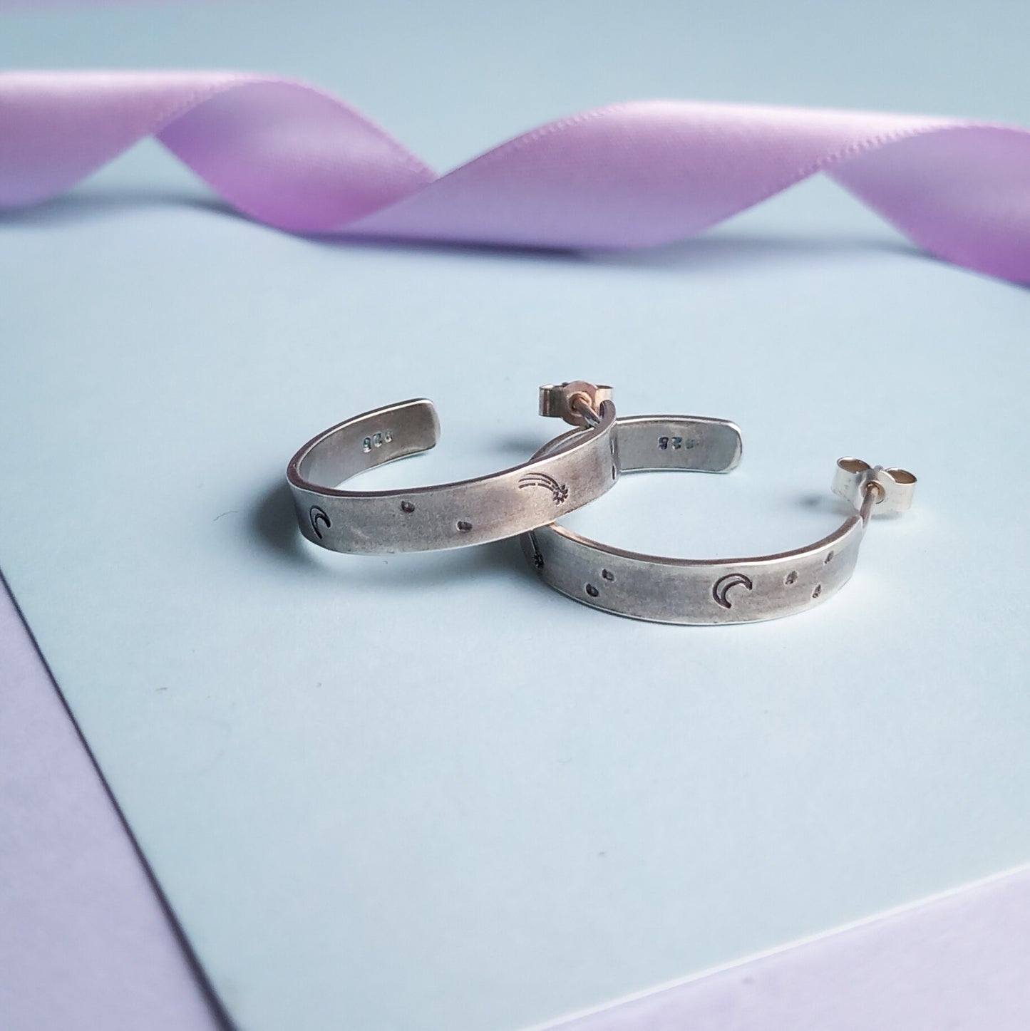 Two recycled silver chunky hoop earrings featuring stamped designs of a moon, comet and stars, elegantly displayed against a pastel background. Styled with a pastel lilac coloured ribbon in the background.
