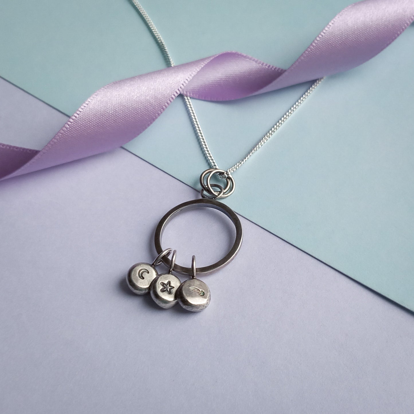 Handmade recycled sterling silver pendant featuring a circular ring with three hanging charms, stamped with a crescent moon, a star, and a shooting star design. The pendant is displayed on a delicate silver chain, styled against a pastel background with a soft lavender ribbon accent.