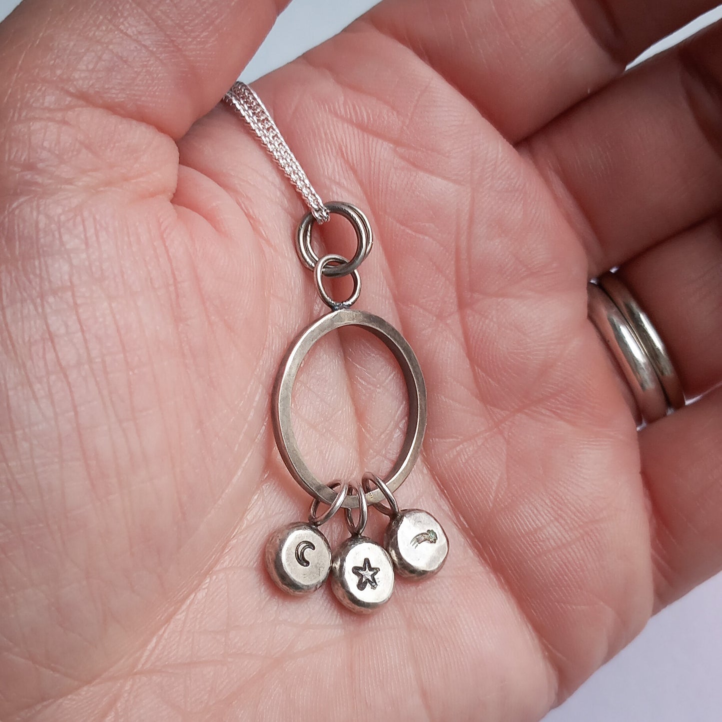A close-up view of a handmade recycled sterling silver pendant, held in the palm of a hand. The pendant features a circular ring with three small charms stamped with designs of a crescent moon, a star, and a shooting star. The piece hangs from a delicate silver chain.