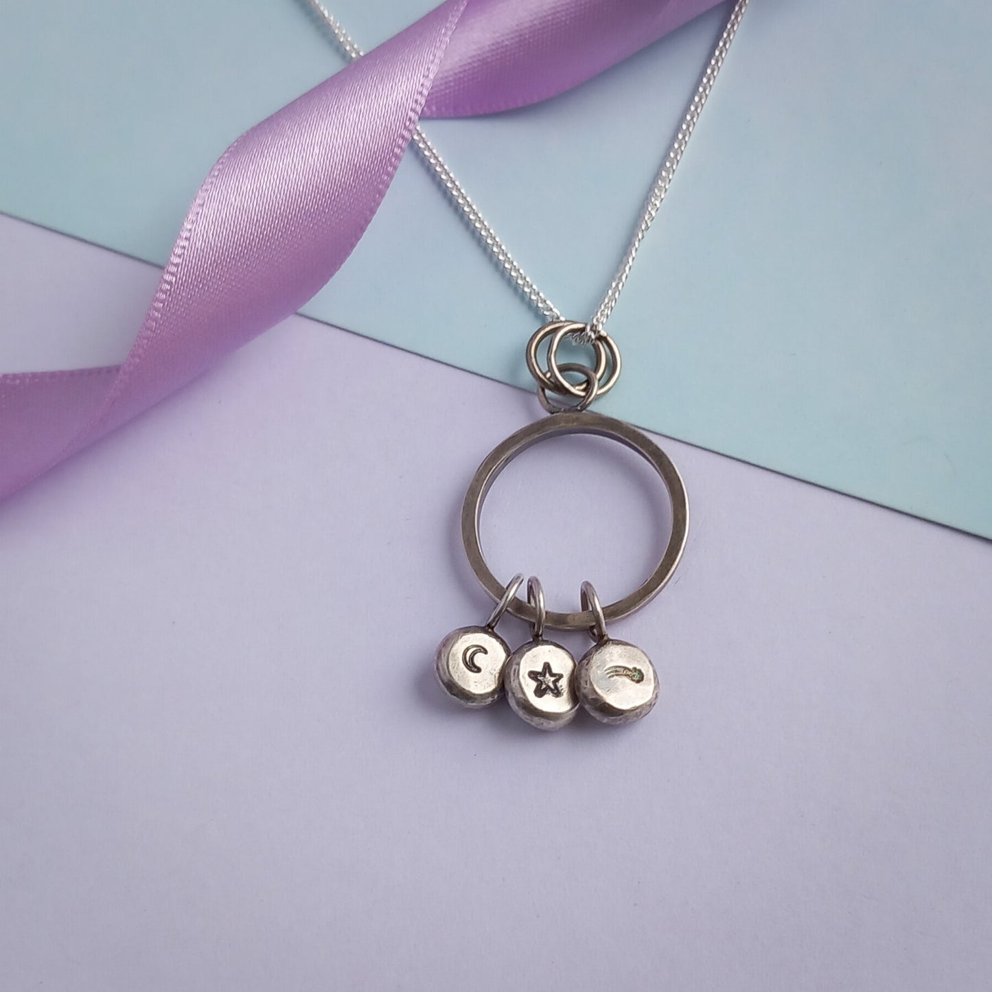 Handmade recycled sterling silver pendant featuring a circular ring with three hanging charms, stamped with a crescent moon, a star, and a shooting star design. The pendant is displayed on a delicate silver chain, styled against a pastel background with a soft lavender ribbon accent.