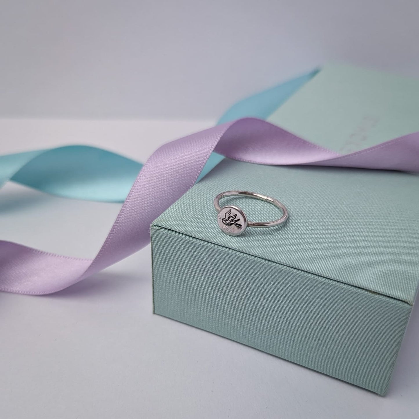 Elegant sterling silver swallow bird ring with a hand stamped disc, displayed on a pastel gift box with lilac and blue satin ribbons. A minimalist and nature-inspired jewellery piece, perfect for gifts and everyday wear.