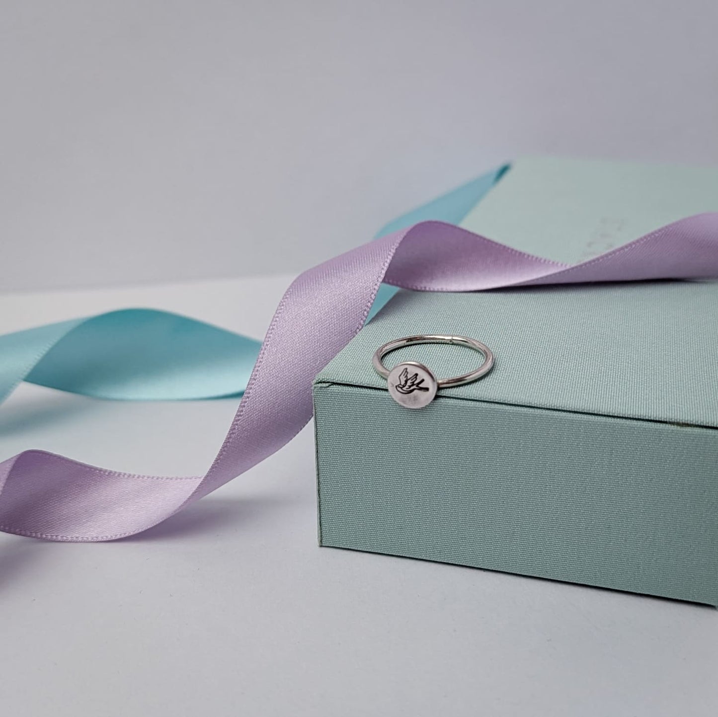 Elegant sterling silver swallow bird ring with a hand stamped disc, displayed on a pastel gift box with lilac and blue satin ribbons. A minimalist and nature-inspired jewellery piece, perfect for gifts and everyday wear.
