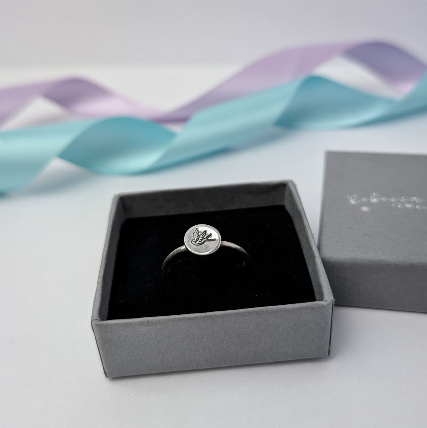 Elegant sterling silver swallow bird ring with a hand stamped disc, displayed in a slate grey ring box with lilac and blue satin ribbons. A minimalist and nature-inspired jewellery piece, perfect for gifts and everyday wear.