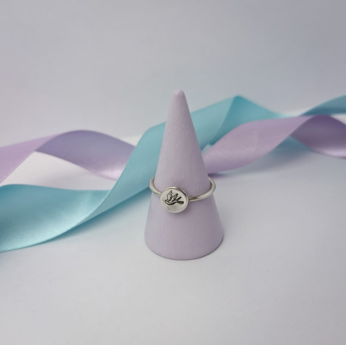 Elegant sterling silver swallow bird ring with a hand stamped disc, displayed on a pastel ring holder with lilac and blue satin ribbons. A minimalist and nature-inspired jewellery piece, perfect for gifts and everyday wear.