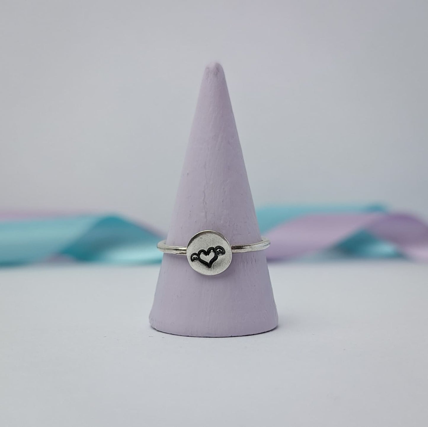 Image shows a simple silver ring with a small disc that features a winged heart design stamped in the centre. The ring is shown displayed on a pale lilac ring holder while the background features pastel blue ribbon, adding a touch of colour.