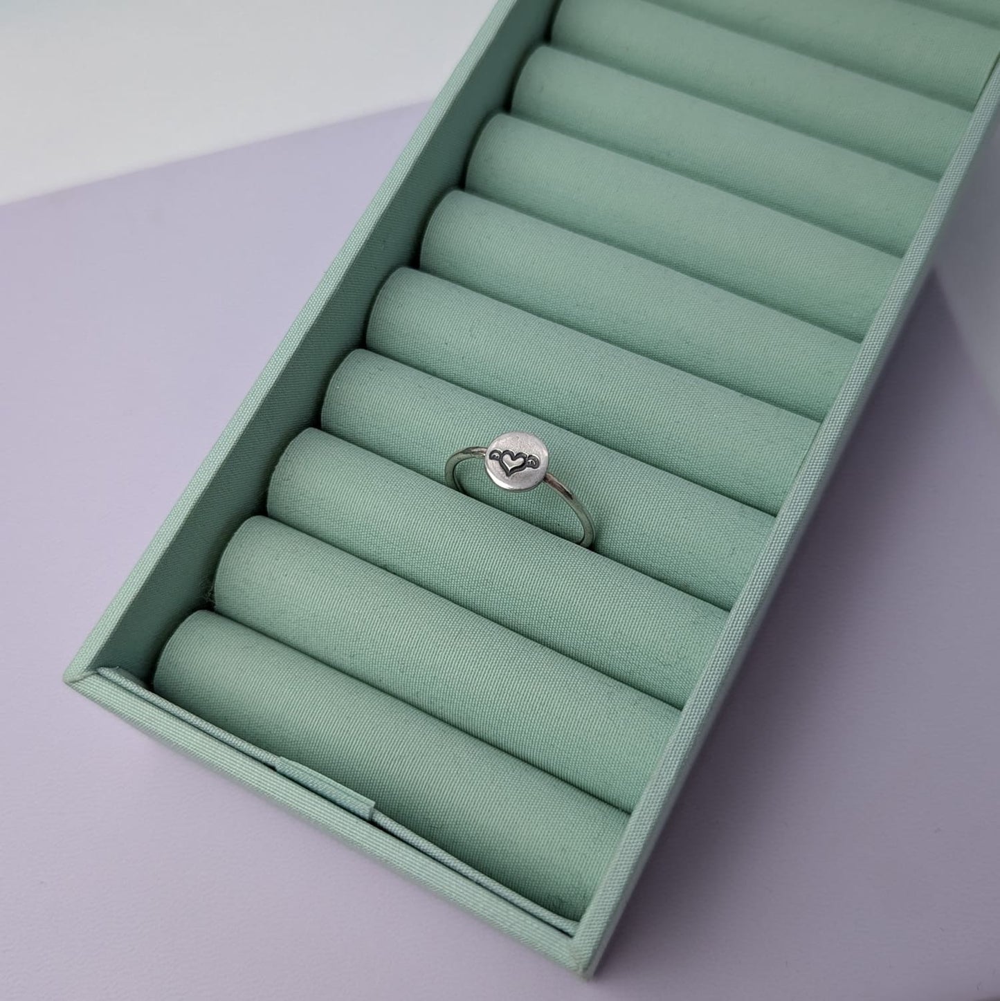 Image shows a simple silver ring with a small disc that features a winged heart design stamped in the centre. The ring sits in a pastel green ring tray with a soft lavender background.