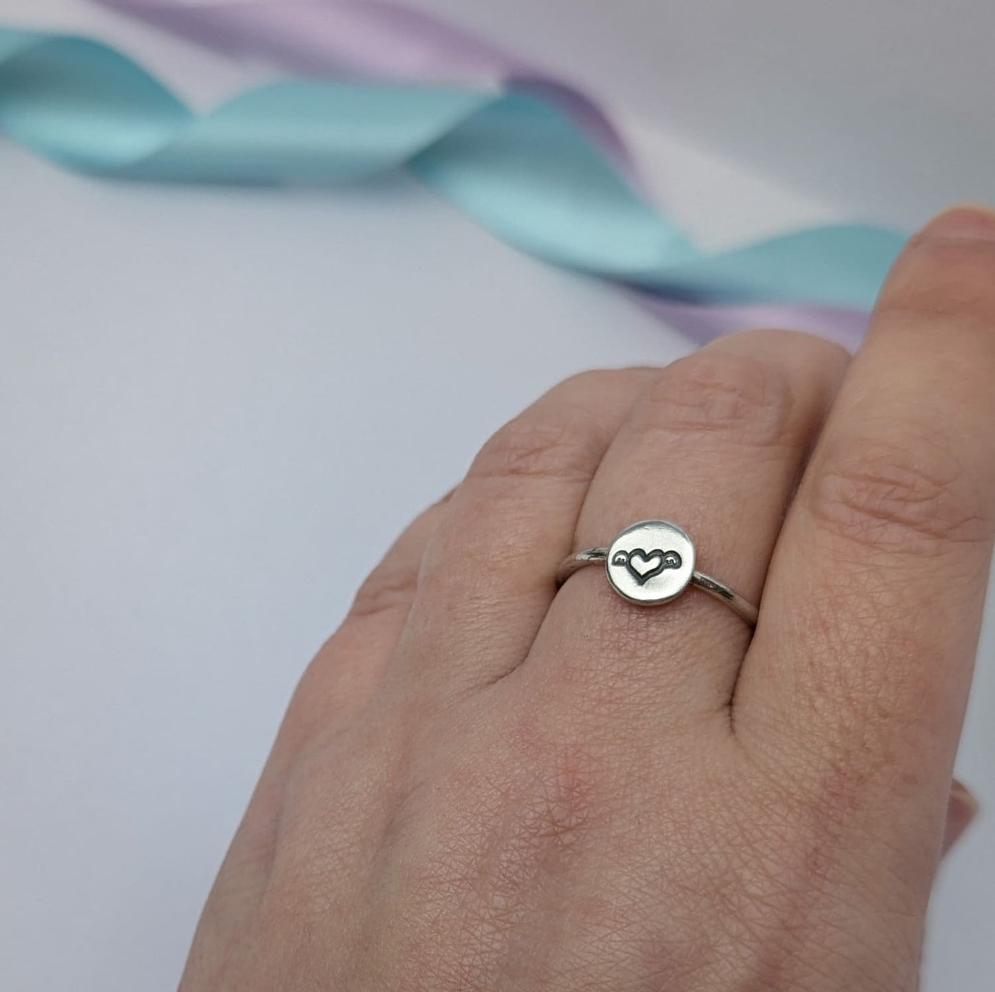 Image shows a simple silver ring with a small disc that features a winged heart design stamped in the centre. The ring is shown being worn on a woman's hand. The background features pastel lavender and blue ribbons which add a touch of colour.