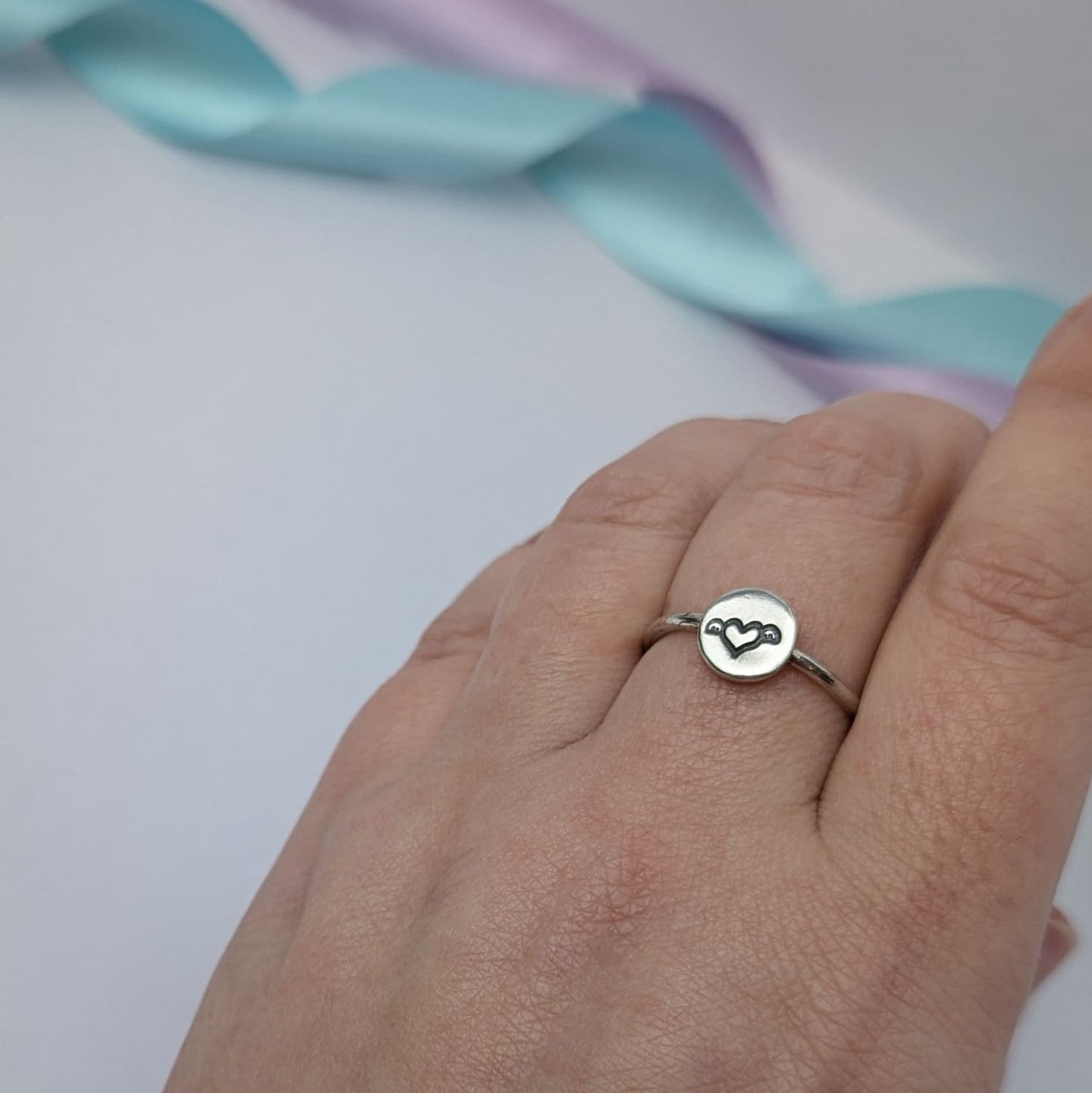 Image shows a simple silver ring with a small disc that features a winged heart design stamped in the centre. The ring is shown being worn on a woman's hand. The background features pastel lavender and blue ribbons which add a touch of colour.