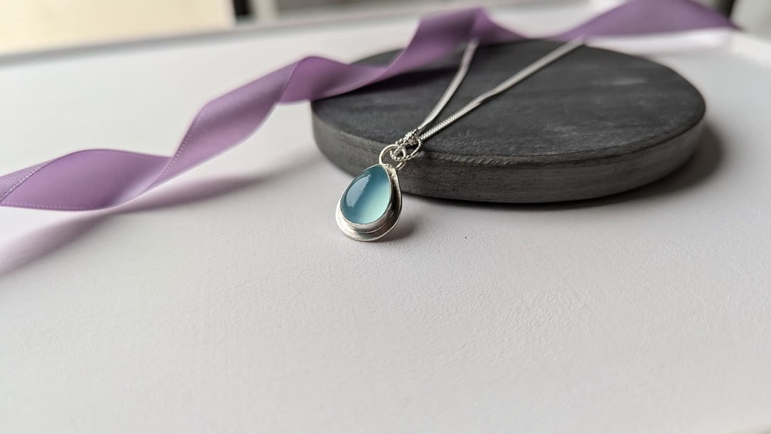 Image of a teardrop shaped silver pendant with a pale blue topaz. The piece is displayed on a slate disc with a lavender ribbon draped behind it, adding a touch of colour.