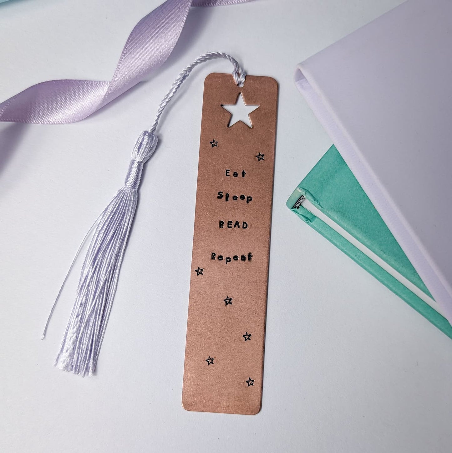Handmade copper bookmark with "Eat Sleep Read Repeat" hand-stamped in black lettering, featuring a star cut out at the top and small stamped stars throughout. A light purple tassel is attached, resting beside a small stack of books. Perfect literary gift for book lovers and avid readers