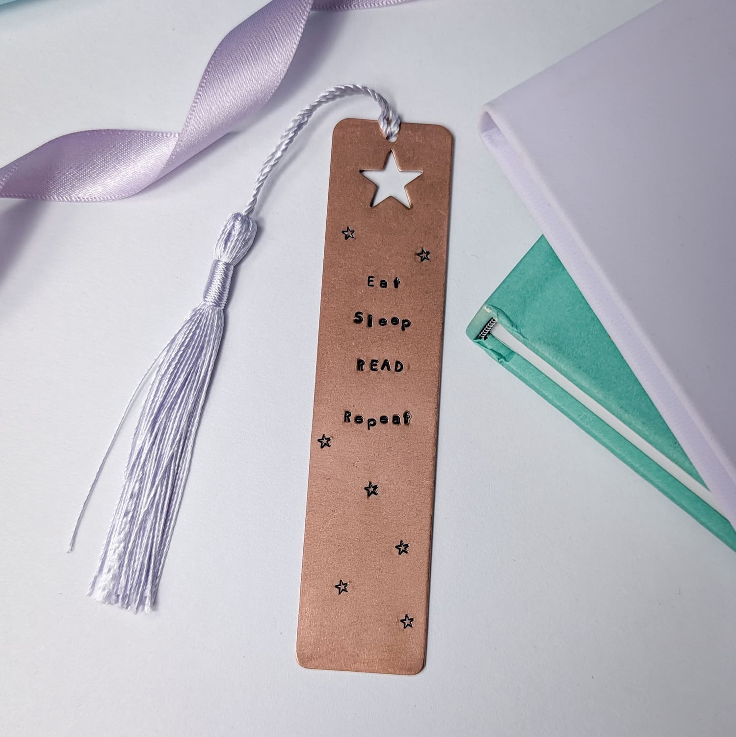 Handmade copper bookmark with "Eat Sleep Read Repeat" hand-stamped in black lettering, featuring a star cut out at the top and small stamped stars throughout. A light purple tassel is attached, resting beside a small stack of books. Perfect literary gift for book lovers and avid readers