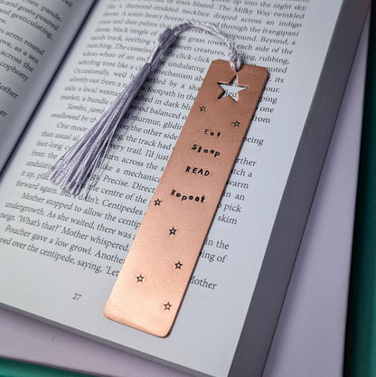 Handmade copper bookmark with "Eat Sleep Read Repeat" hand-stamped in black lettering, featuring a star cut out at the top and small stamped stars throughout. A light purple tassel is attached, resting on an open book with printed text. Perfect literary gift for book lovers and avid readers