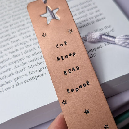 Handmade copper bookmark with "Eat Sleep Read Repeat" hand-stamped in black lettering, featuring a star cut out at the top and small stamped stars throughout. A light purple tassel is attached, resting on an open book with printed text. Perfect literary gift for book lovers and avid readers