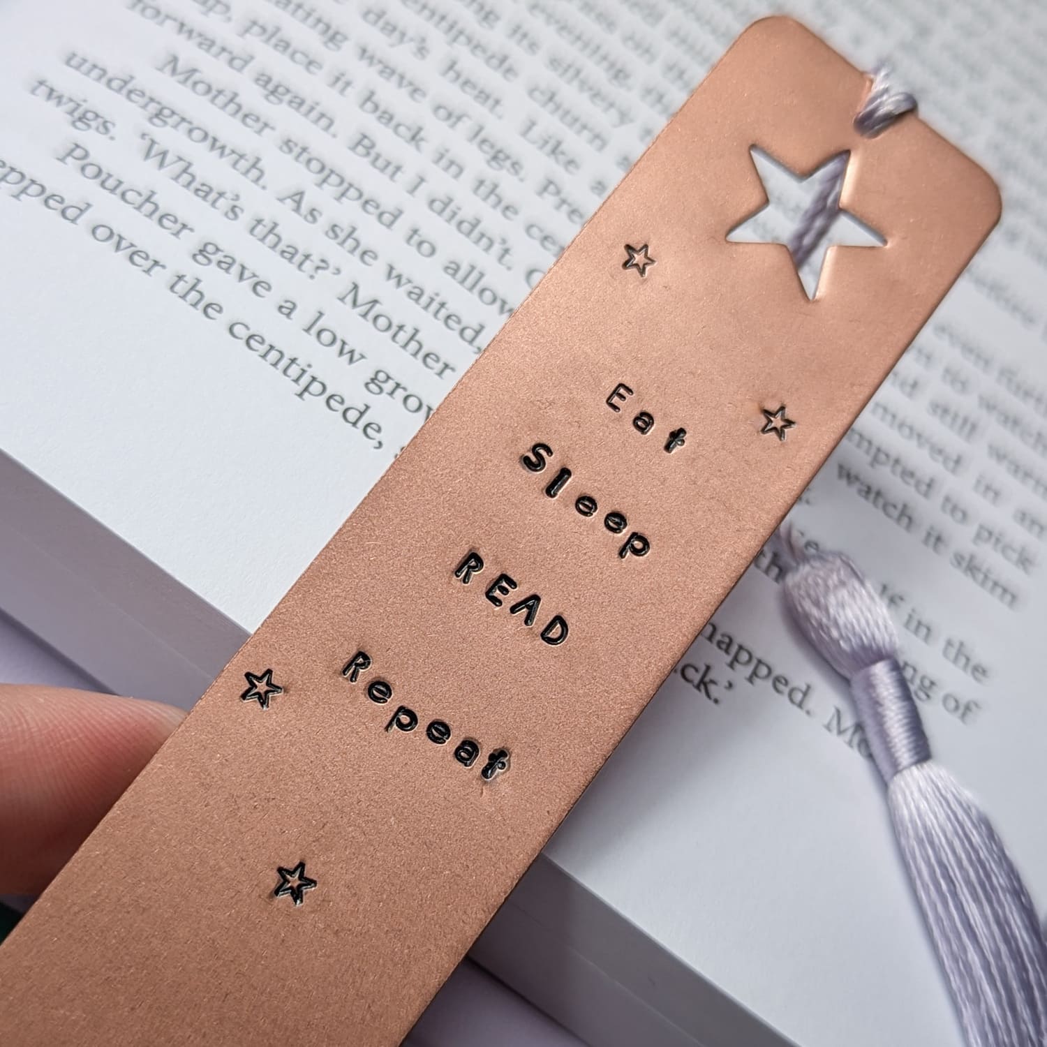 Handmade copper bookmark with "Eat Sleep Read Repeat" hand-stamped in black lettering, featuring a star cut out at the top and small stamped stars throughout. A light purple tassel is attached, resting on an open book with printed text. Perfect literary gift for book lovers and avid readers
