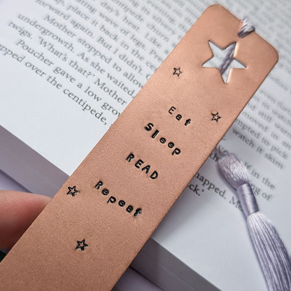 Handmade copper bookmark with "Eat Sleep Read Repeat" hand-stamped in black lettering, featuring a star cut out at the top and small stamped stars throughout. A light purple tassel is attached, resting on an open book with printed text. Perfect literary gift for book lovers and avid readers