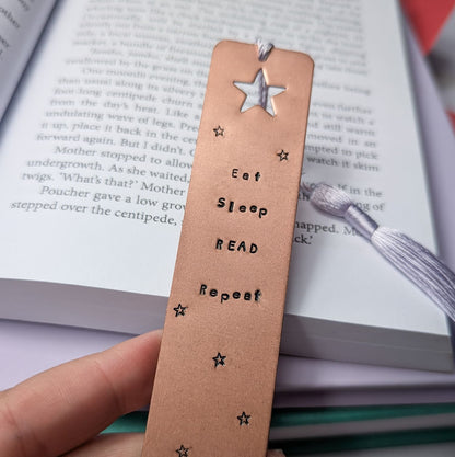 Handmade copper bookmark with "Eat Sleep Read Repeat" hand-stamped in black lettering, featuring a star cutout at the top and small engraved stars throughout. A light purple tassel is attached, resting on an open book with printed text. Perfect literary gift for book lovers and avid readers