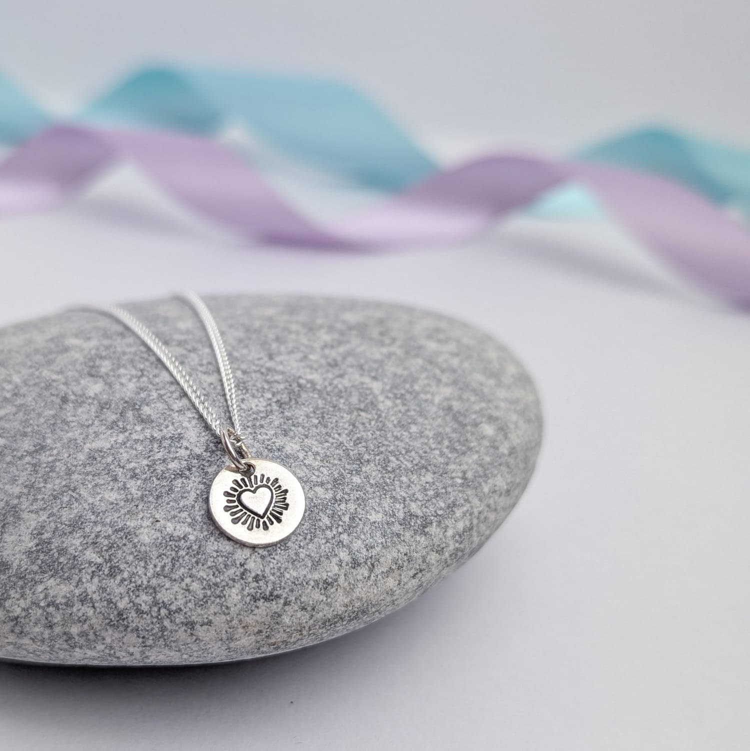 Photograph of a delicate silver pendant necklace resting on a smooth grey stone. The round disc pendant features a hand-stamped heart in the centre, surrounded by radiating lines resembling sun rays. The necklace has a fine silver chain. The background is softly blurred, with pastel blue and lavender ribbons adding a touch of colour.