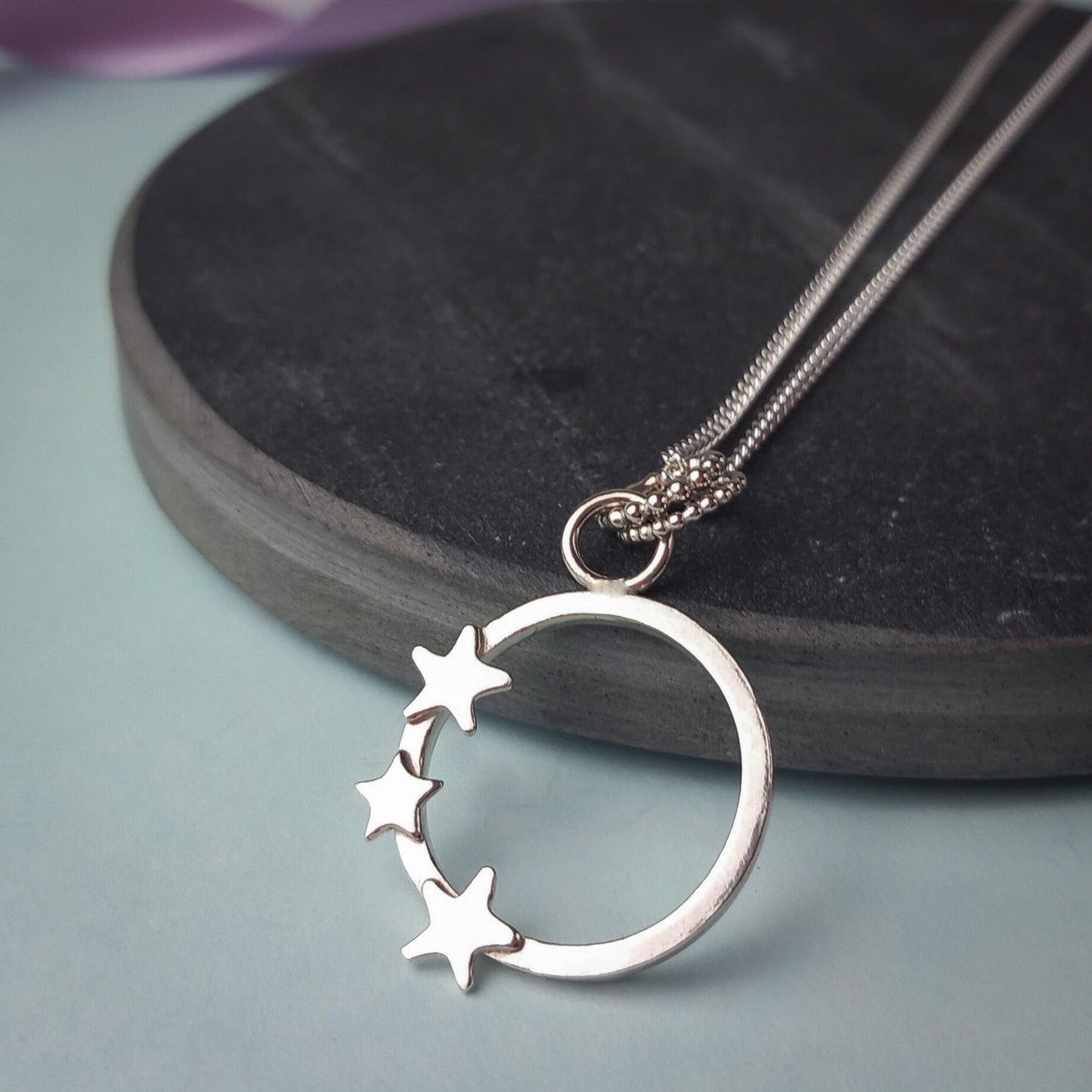 Elegant sterling silver pendant necklace featuring an open circle design adorned with three small star accents. The delicate pendant hangs from a fine silver chain, displayed on a dark stone background. A minimalist celestial jewellery piece, perfect for everyday wear or as a thoughtful gift