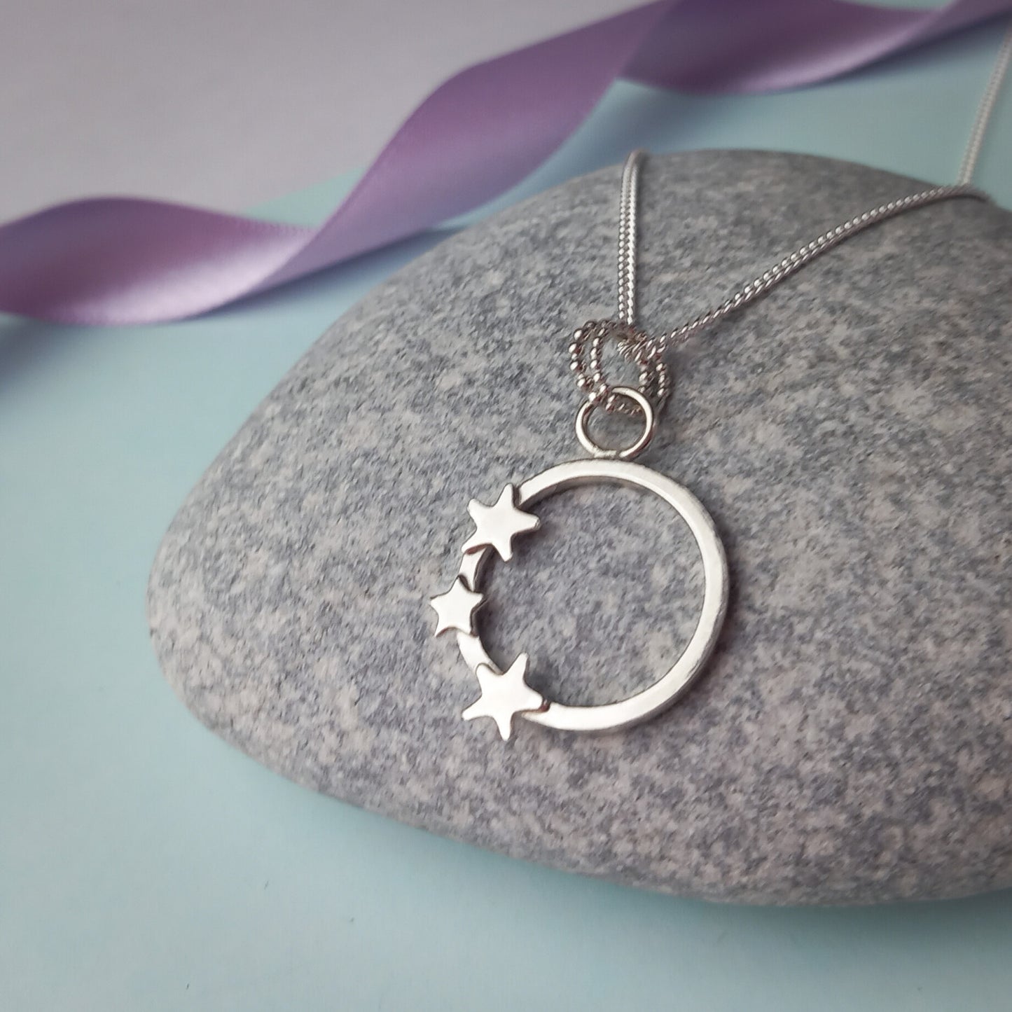Elegant sterling silver pendant necklace featuring an open circle design adorned with three small star accents. The delicate pendant hangs from a fine silver chain, displayed on a stone background. A minimalist celestial jewellery piece, perfect for everyday wear or as a thoughtful gift