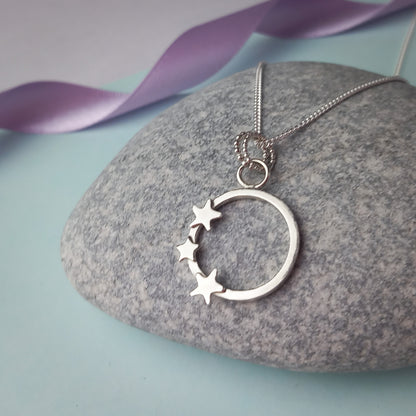 Elegant sterling silver pendant necklace featuring an open circle design adorned with three small star accents. The delicate pendant hangs from a fine silver chain, displayed on a stone background. A minimalist celestial jewellery piece, perfect for everyday wear or as a thoughtful gift