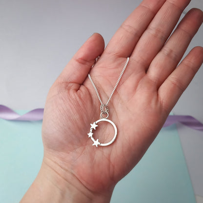 Elegant sterling silver pendant necklace featuring an open circle design adorned with three small star accents. The delicate pendant hangs from a fine silver chain, displayed in the palm of a hand. A minimalist celestial jewellery piece, perfect for everyday wear or as a thoughtful gift