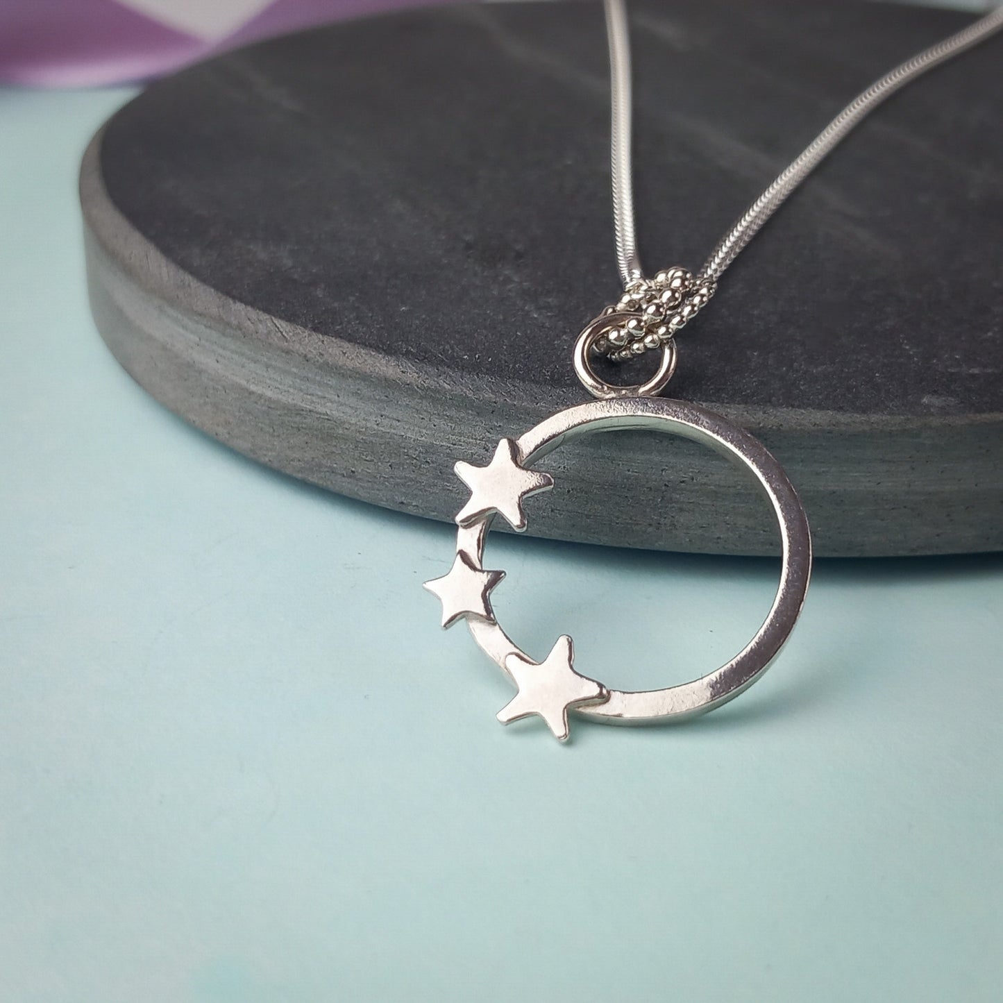 Elegant sterling silver pendant necklace featuring an open circle design adorned with three small star accents. The delicate pendant hangs from a fine silver chain, displayed on a dark stone background. A minimalist celestial jewellery piece, perfect for everyday wear or as a thoughtful gift