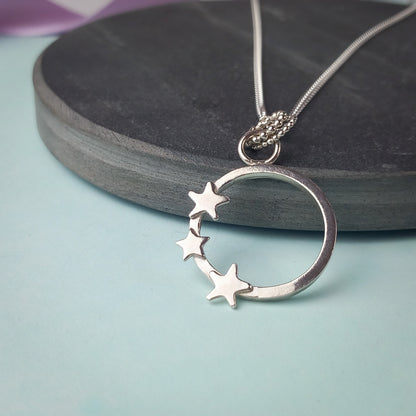 Elegant sterling silver pendant necklace featuring an open circle design adorned with three small star accents. The delicate pendant hangs from a fine silver chain, displayed on a dark stone background. A minimalist celestial jewellery piece, perfect for everyday wear or as a thoughtful gift