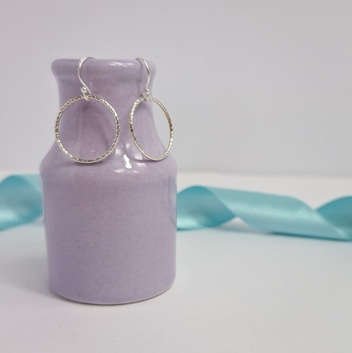 Photograph of a pair of silver open circle earrings hanging from the edge of a tiny purple vase. Each earring features an open circle with a soft hammered texture that beautifully catches the light. The background is softly blurred, with pastel blue ribbons adding a touch of colour.