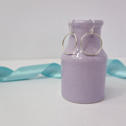 Photograph of a pair of silver open circle earrings hanging from the edge of a tiny purple vase. Each earring features an open circle with a soft hammered texture that beautifully catches the light. The background is softly blurred, with pastel blue ribbons adding a touch of colour.
