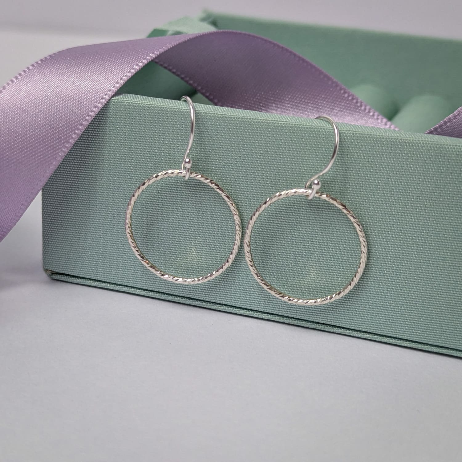 Photograph of a pair of silver open circle earrings resting on a pastel green ring box. Each earring features an open circle with a soft hammered texture that beautifully catches the light. The background is softly blurred, with pastel lavender ribbons adding a touch of colour.