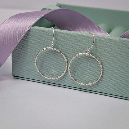 Photograph of a pair of silver open circle earrings resting on a pastel green ring box. Each earring features an open circle with a soft hammered texture that beautifully catches the light. The background is softly blurred, with pastel lavender ribbons adding a touch of colour.
