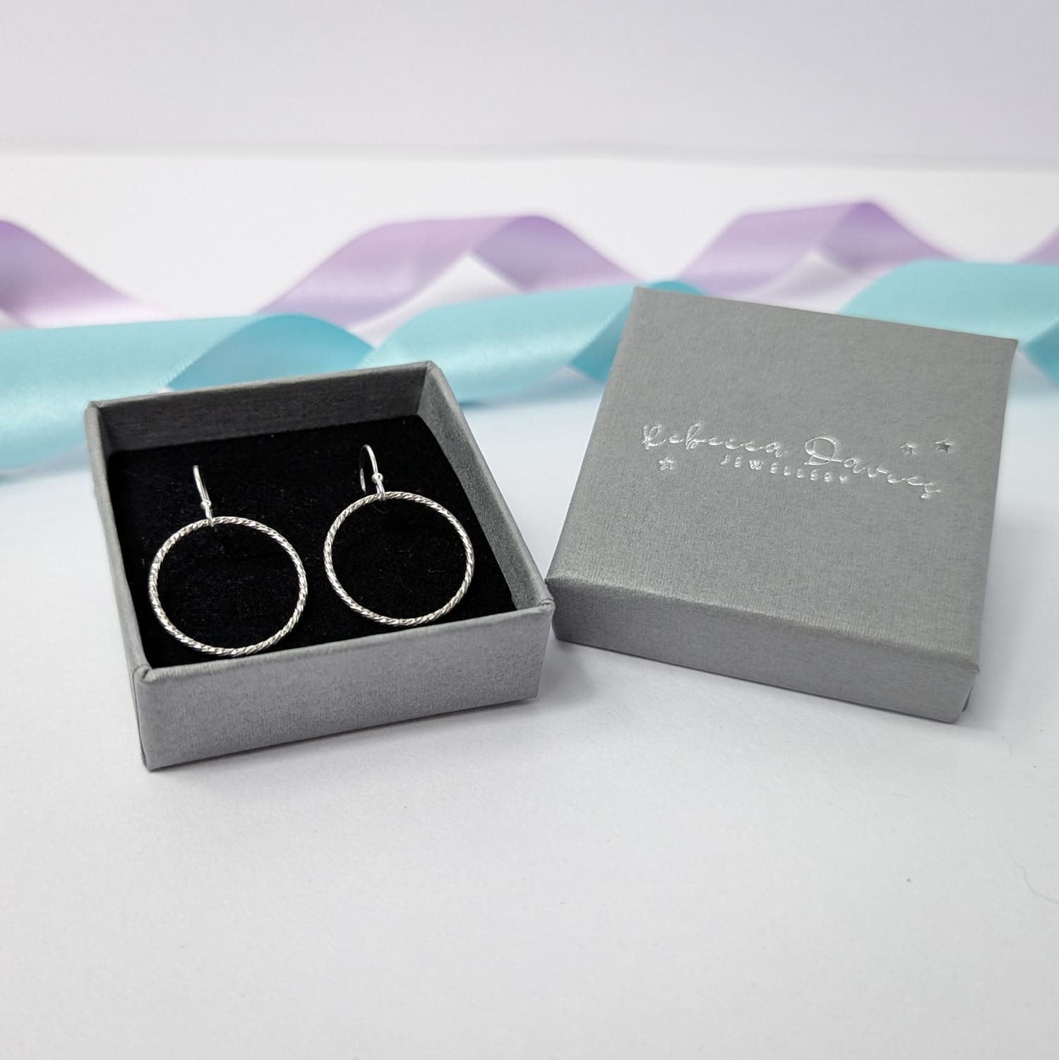 Photograph of a pair of silver open circle earrings displayed in a luxury grey gift box. Each earring features an open circle with a soft hammered texture that beautifully catches the light. The background is softly blurred, with pastel blue and lavender ribbons adding a touch of colour.