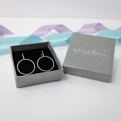 Photograph of a pair of silver open circle earrings displayed in a luxury grey gift box. Each earring features an open circle with a soft hammered texture that beautifully catches the light. The background is softly blurred, with pastel blue and lavender ribbons adding a touch of colour.