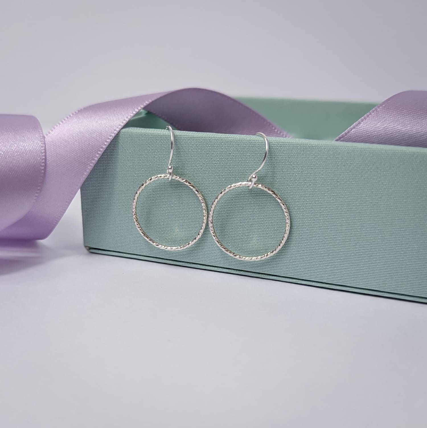 Photograph of a pair of silver open circle earrings resting on a pastel green ring box. Each earring features an open circle with a soft hammered texture that beautifully catches the light. The background is softly blurred, with pastel lavender ribbons adding a touch of colour.