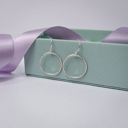 Photograph of a pair of silver open circle earrings resting on a pastel green ring box. Each earring features an open circle with a soft hammered texture that beautifully catches the light. The background is softly blurred, with pastel lavender ribbons adding a touch of colour.