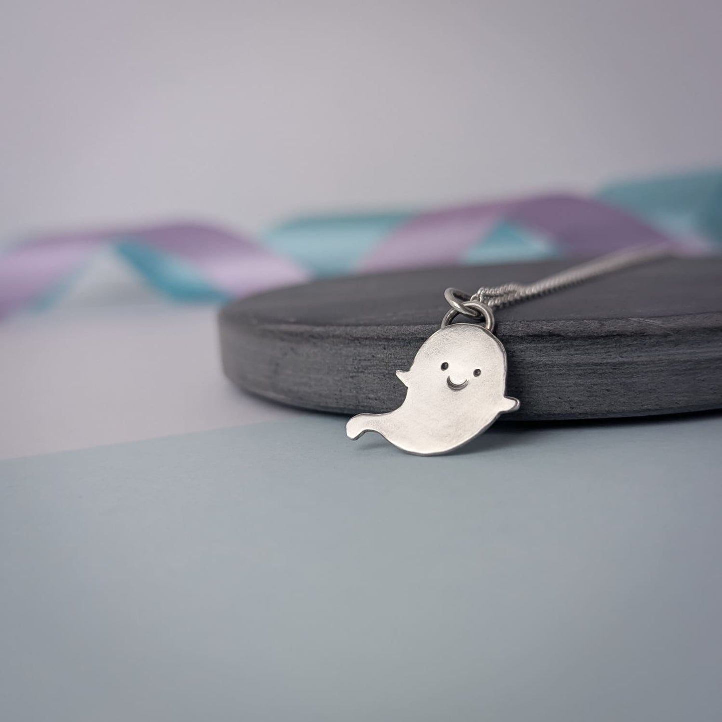 Adorable handmade sterling silver ghost necklace. The pendant is shown on a chain against a soft pastel background.