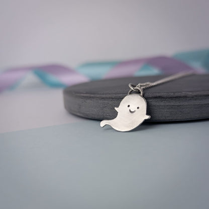 Adorable handmade sterling silver ghost necklace. The pendant is shown on a chain against a soft pastel background.