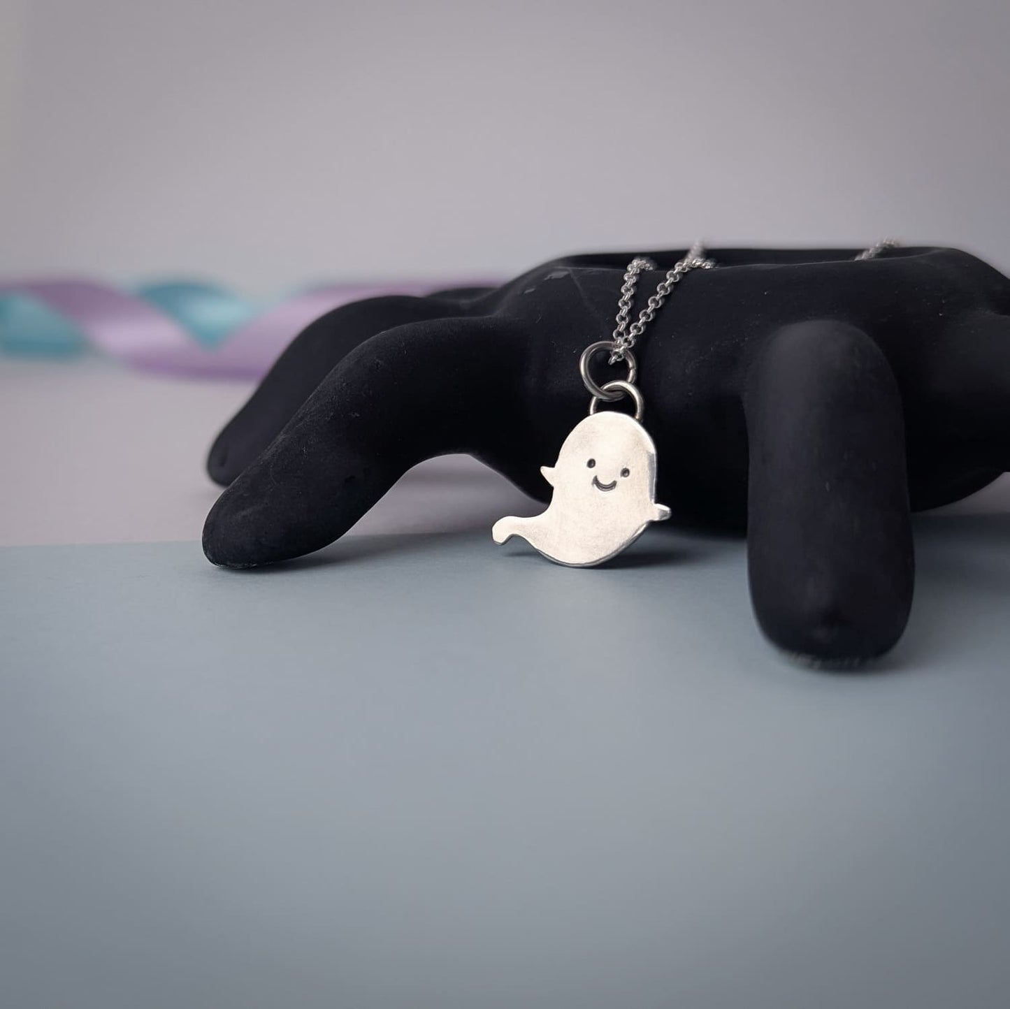 Ghost-shaped pendant on a chain displayed on a black spider against a soft pastel background.