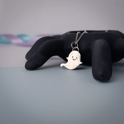 Ghost-shaped pendant on a chain displayed on a black spider against a soft pastel background.