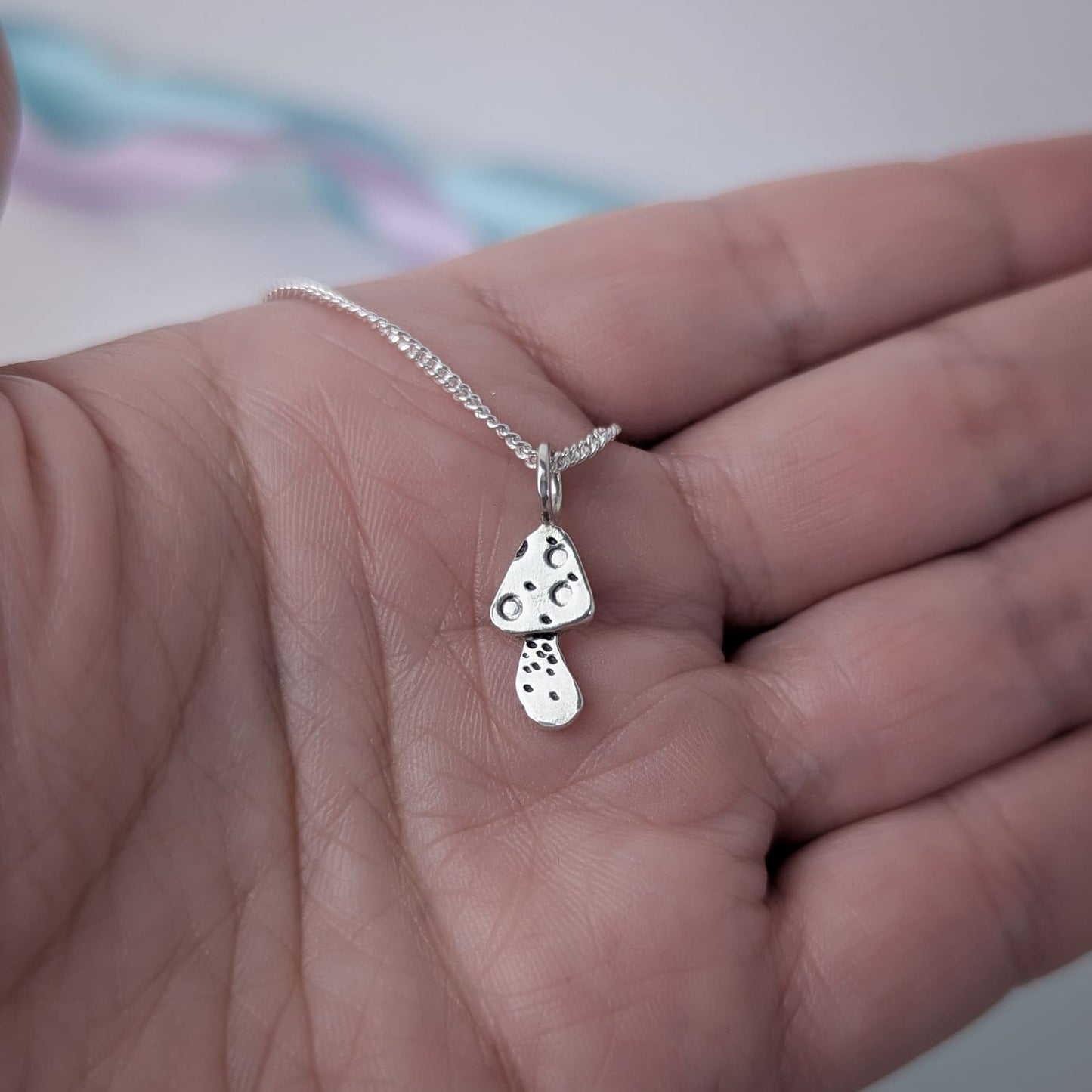 Tiny handmade sterling silver necklace with a mushroom pendant held in a hand against a blurred pastel background