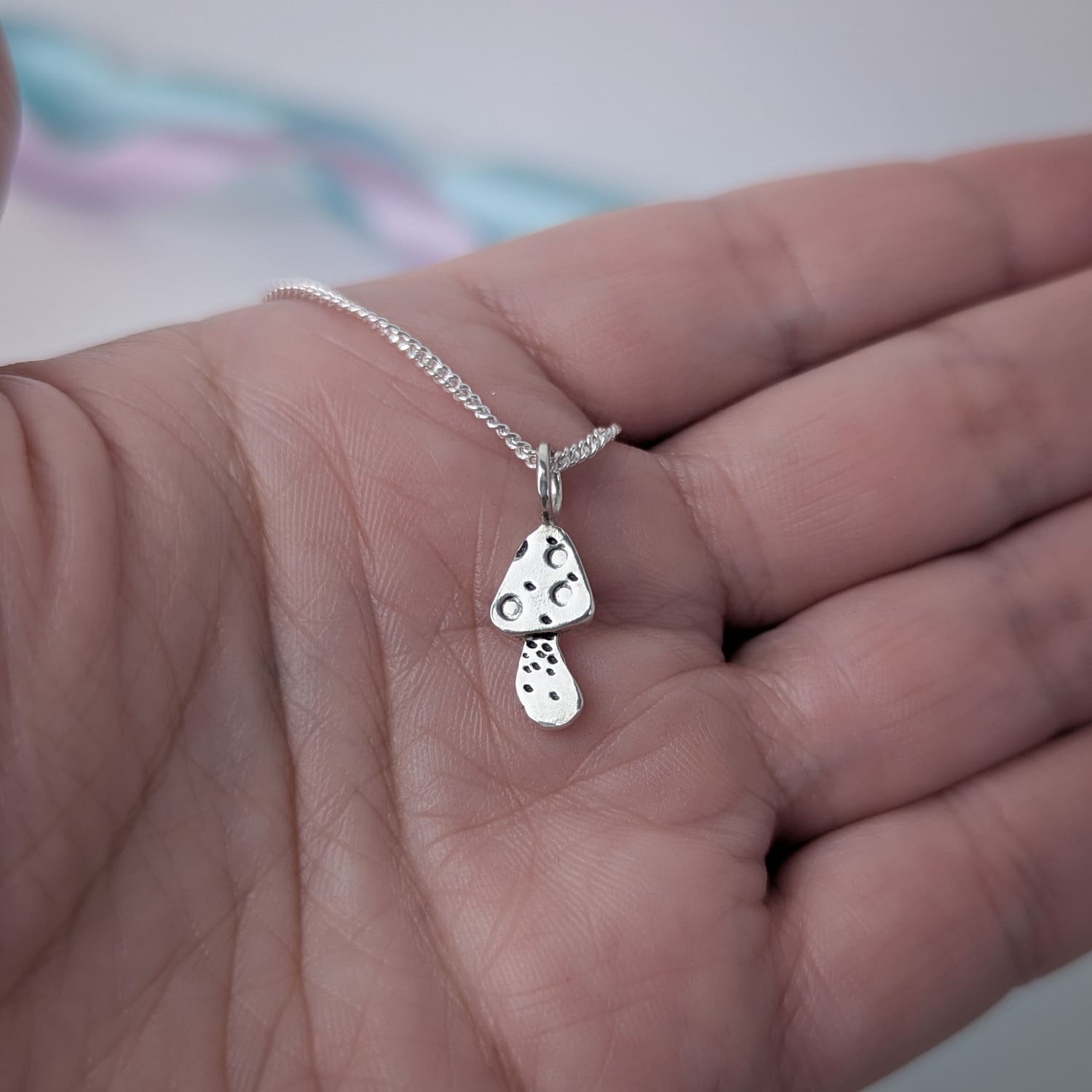 Tiny handmade sterling silver necklace with a mushroom pendant held in a hand against a blurred pastel background